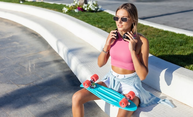 Jovem loira vestida com roupas elegantes e óculos escuros com skate e fones de ouvido ao ar livre em um dia ensolarado de verão