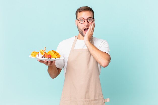 Jovem loiro bonito se sentindo chocado e com medo. cozinhar waffles conceito