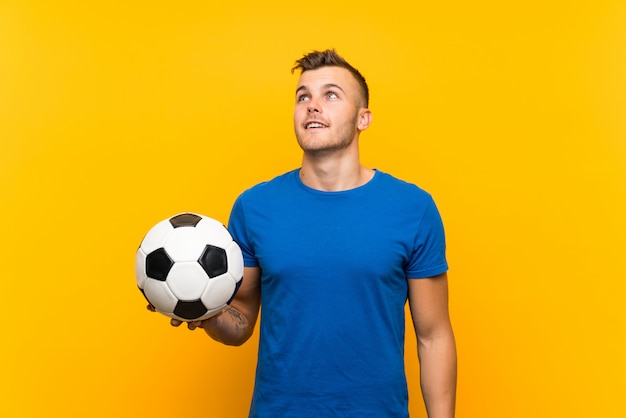 Jovem loiro bonito segurando uma bola de futebol sobre parede amarela isolada, olhando para cima enquanto sorrindo