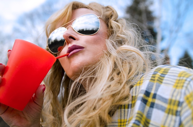 Foto jovem, loiro, mulher, com, óculos de sol, bebendo, ligado, palha, de, vermelho, vidro