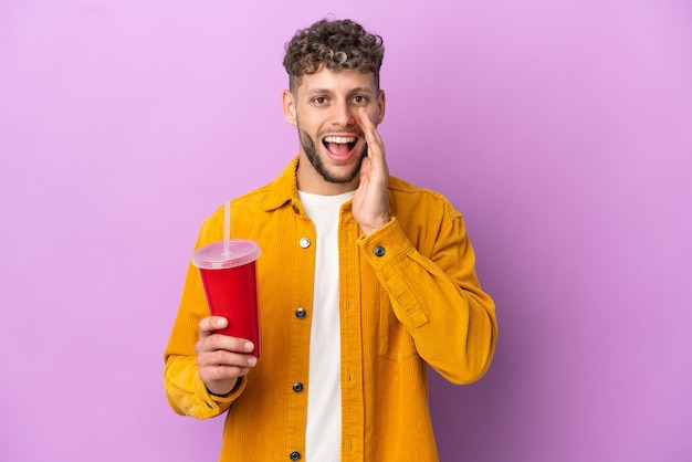 Jovem loiro segurando refrigerante isolado em um fundo roxo e gritando com a boca bem aberta