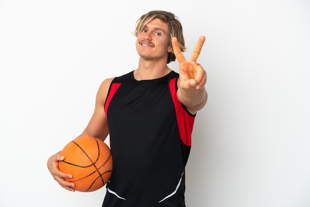 Jovem loiro segurando uma bola de basquete isolada na parede branca sorrindo e mostrando o sinal da vitória