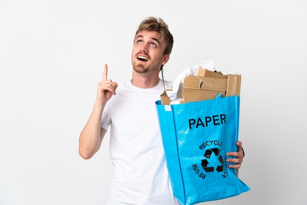 Jovem loiro segurando uma sacola de reciclagem cheia de papel para reciclar isolado no fundo branco apontando para cima e surpreso