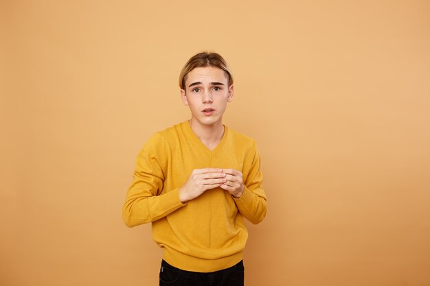 Jovem loiro vestido com um suéter amarelo está posando no estúdio sobre o fundo bege.