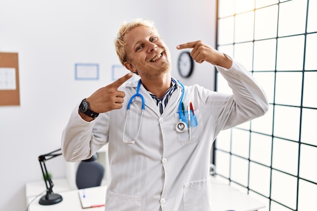 Jovem loiro vestindo uniforme médico e estetoscópio na clínica sorrindo alegre mostrando e apontando com os dedos, dentes e boca. conceito de saúde bucal.