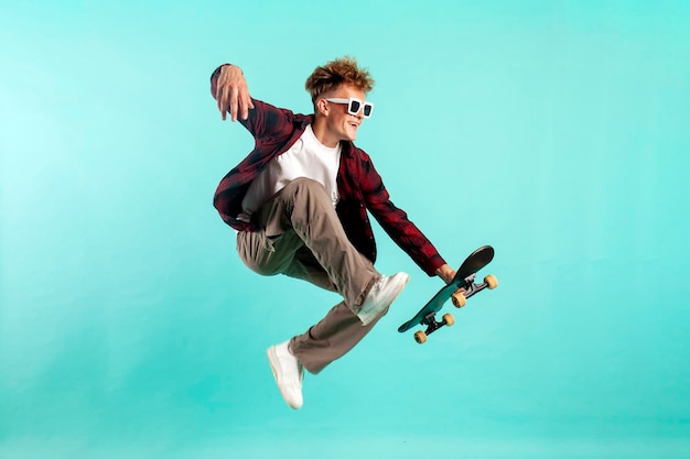 Jovem louco anda de skate e salta em fundo azul isolado hipster em óculos de sol voa