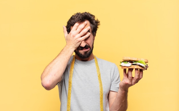 Jovem louco barbudo com uma expressão triste em uma dieta e segurando um sanduíche