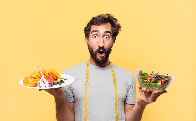 Jovem louco barbudo fazendo dieta duvidosa ou expressão incerta, segurando uma salada e waffles