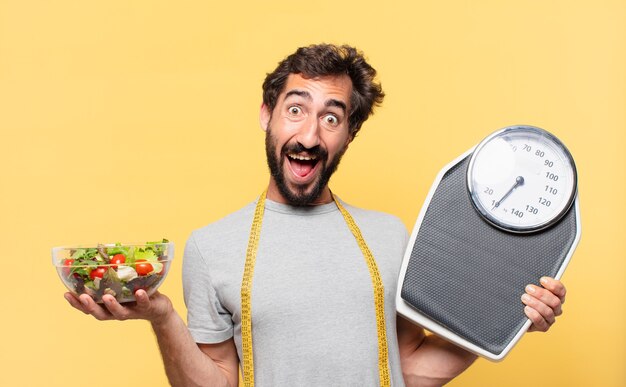 Jovem louco barbudo fazendo dieta expressão de surpresa e segurando uma balança e uma salada