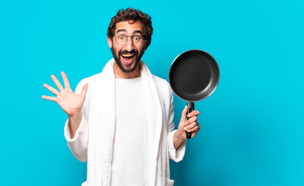 Jovem louco barbudo vestindo um roupão de banho cozinhando com uma panela