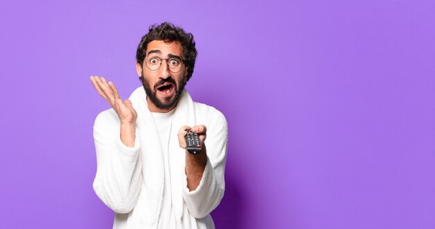 Jovem louco de barba usando roupão de banho e controle remoto da televisão