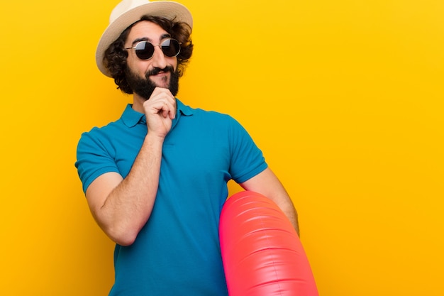 Jovem louco sorrindo com uma expressão confiante feliz com a mão no queixo, pensando e olhando para o lado contra a parede laranja