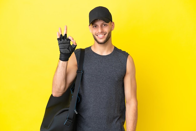 Jovem loura esportiva com bolsa esportiva isolada em fundo amarelo, mostrando sinal de ok com os dedos
