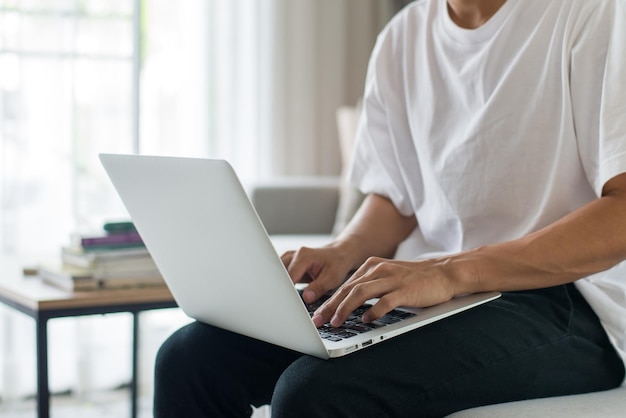 Jovem macho vestindo camiseta branca sentado no sofá e usando o laptop para verificar e-mails e responder a mensagens em casa