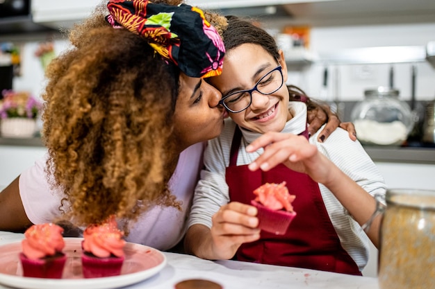 Jovem mãe abraçando e beijando sua filha enquanto cozinha bolos juntos para uma festa