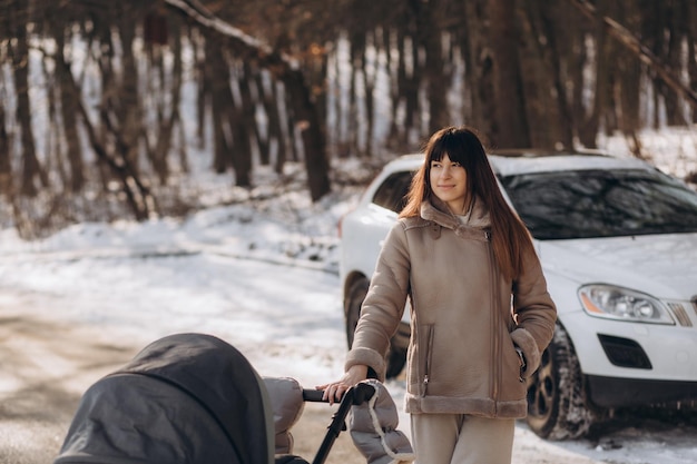 Jovem mãe adulta empurrando o carrinho de bebê e andando na calçada coberta de neve no parque em um dia frio de inverno, passando um tempo com o recém-nascido e respirando ar fresco