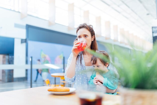 Jovem mãe adulta tomando um refrigerante e comendo algo com seu bebê em um shopping