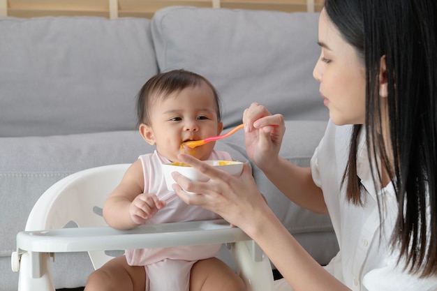 Jovem mãe ajudando o bebê a comer alimentos misturados na cadeira de bebê