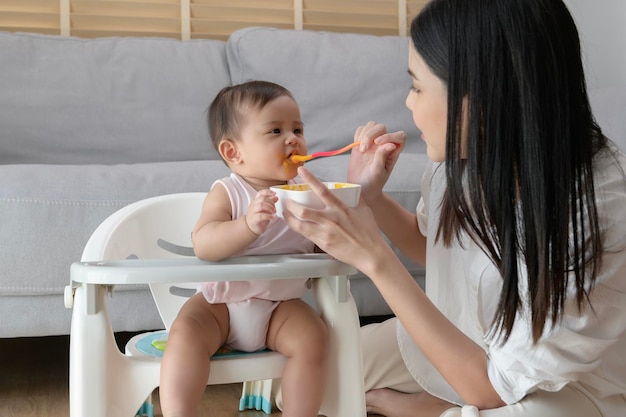 Jovem mãe ajudando o bebê a comer alimentos misturados na cadeira de bebê