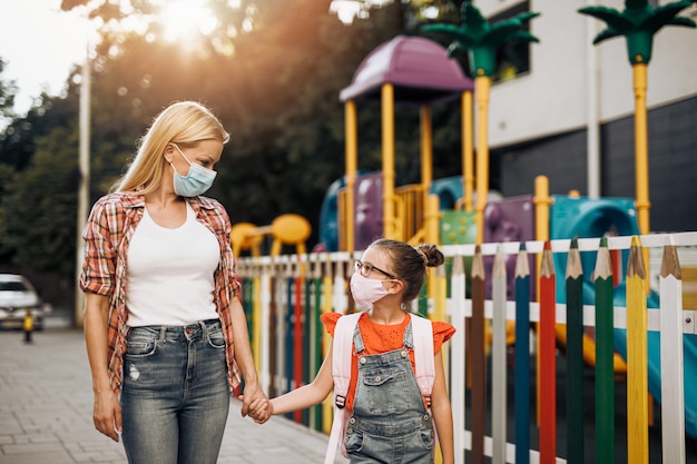 Jovem mãe andando com sua filha na rua da cidade. Eles usam máscaras de proteção facial. De volta às aulas e ao novo conceito de estilo de vida do coronavírus.