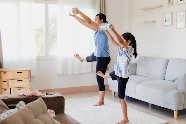 Jovem mãe asiática e a filha dela fazendo alongamento, ioga, exercícios de fitness e usando a garrafa de água para serem halteres juntos em casa na rotina diária.