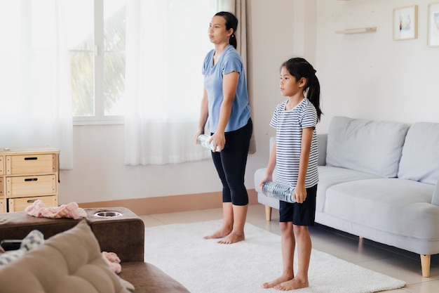 Jovem mãe asiática e a filha dela fazendo alongamento, ioga, exercícios de fitness e usando a garrafa de água para serem halteres juntos em casa na rotina diária.