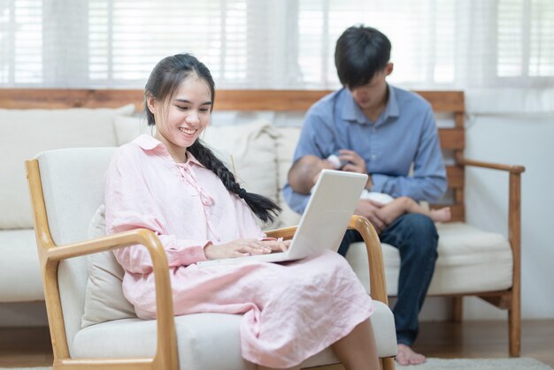 Jovem mãe asiática usando laptop e pai alimentando o bebê