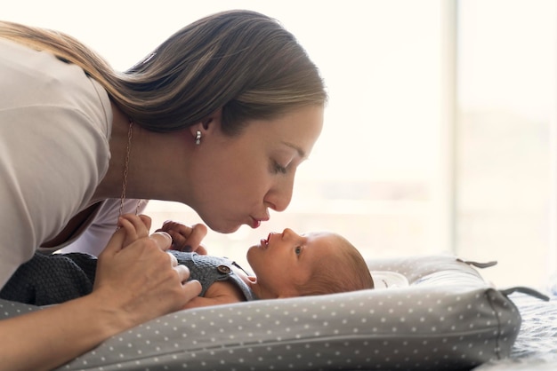 Jovem mãe beijando seu bebê recém-nascido na cama perto da janela