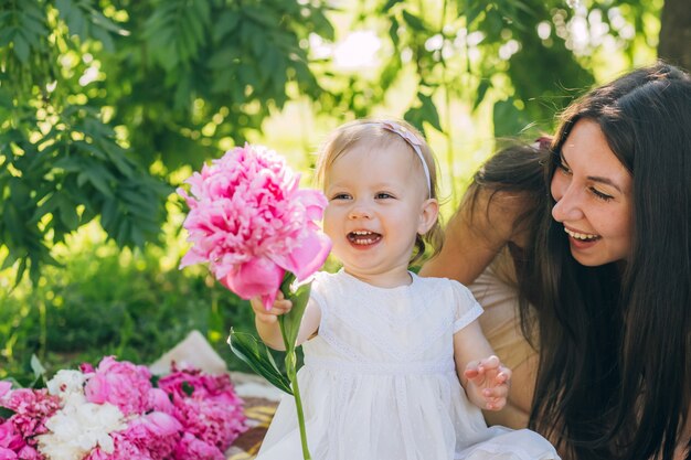 Jovem mãe bonita com filha mentir sobre uma manta no parque. Família recreação ao ar livre. Piquenique em família verão no parque. Mãe e filha ao ar livre, cercada por peônias rosa.