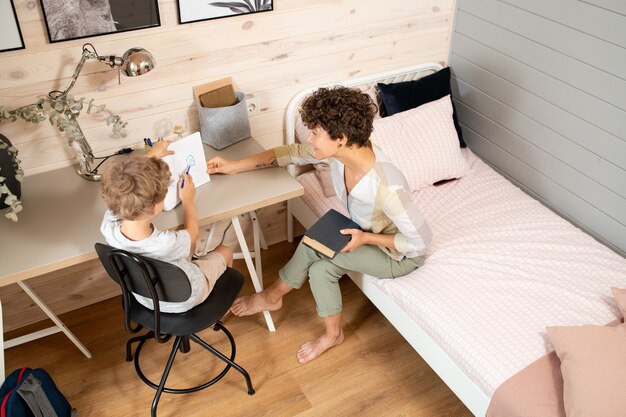 Jovem mãe com o livro sentado na cama ao lado da mesa, onde o filho está sentado e mostrando a foto desenhada no caderno enquanto faz a lição de casa