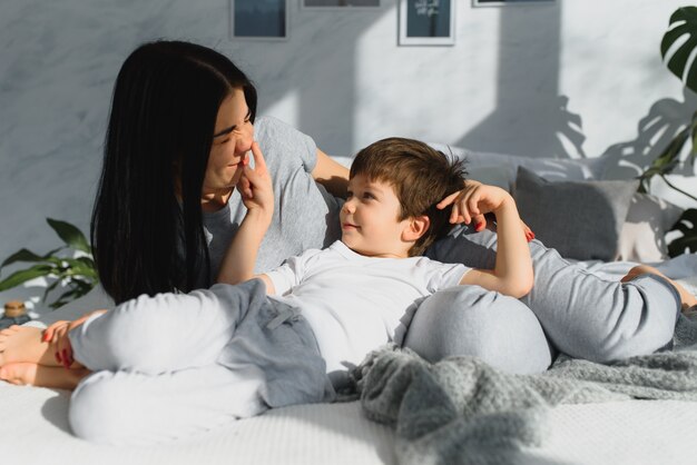 Jovem mãe com seu filho pequeno de 4 anos de pijama relaxando e brincando na cama