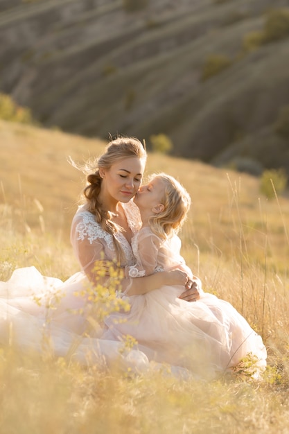 Jovem mãe com uma filha pequena em vestidos cor de rosa estão sentados no campo.