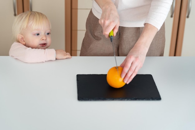 Jovem mãe corta uma laranja para o bebê que está por perto Segurança na cozinha