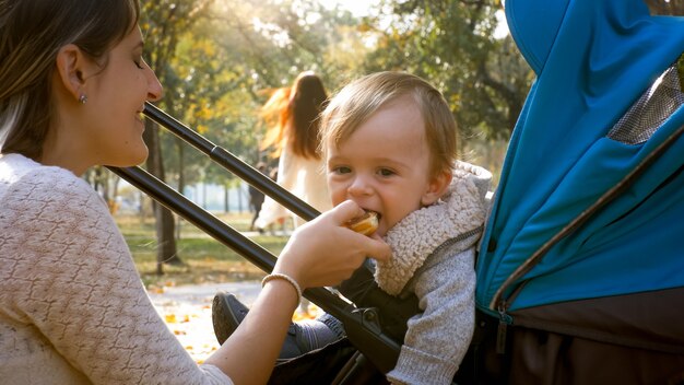 Jovem mãe dando comida para seu filho bebê fofo sentado no carrinho no parque auutmn.