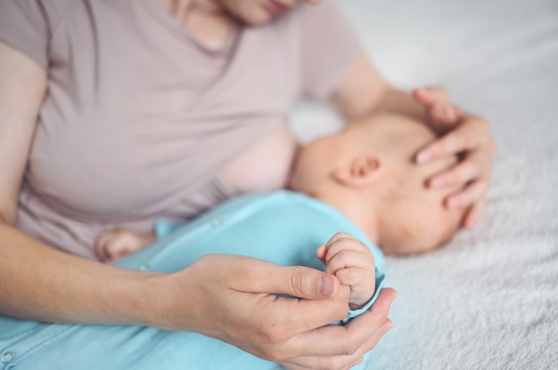 Jovem mãe deita-se com um bebê recém-nascido fofo com macacão azul na cama, abraçando-o e amamentando-o com leite materno