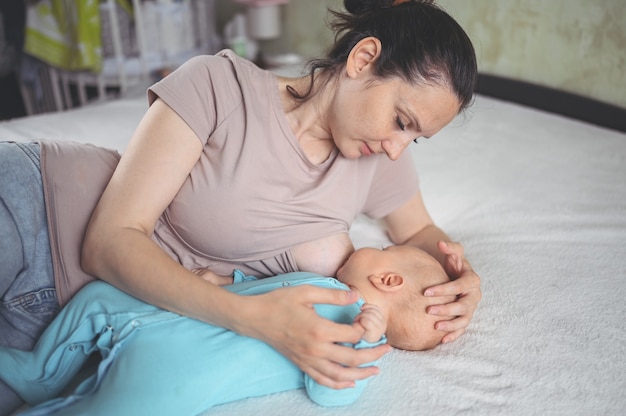 Jovem mãe deita-se com um bebê recém-nascido fofo com macacão azul na cama, abraçando-o e amamentando-o com leite materno