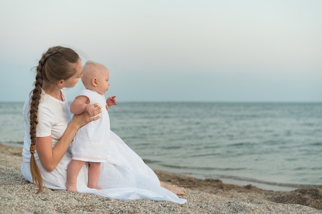 Jovem mãe e bebê estão descansando na praia. Linda mãe segurando bebê na beira-mar