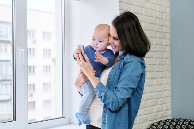 Jovem mãe e filho bebê em seus braços estão olhando para a tela do smartphone, videochamada, comunicação on-line. Família feliz em casa, perto da janela, temporada de outono de inverno