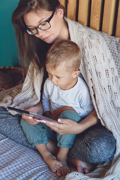 Jovem mãe e filho usando um tablet sob um cobertor aconchegante no quarto de sua casa