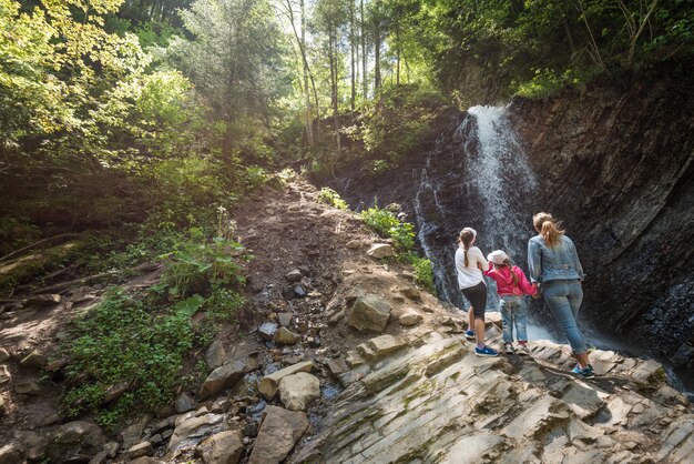 Jovem mãe e filhos caminham pela mata com uma vista deslumbrante da extensão natural das formações rochosas das colinas e de uma cachoeira