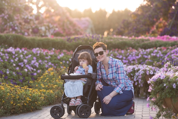 jovem mãe e menina bonitinha em um carrinho de bebê, desfrutando de um lindo dia no jardim de flores