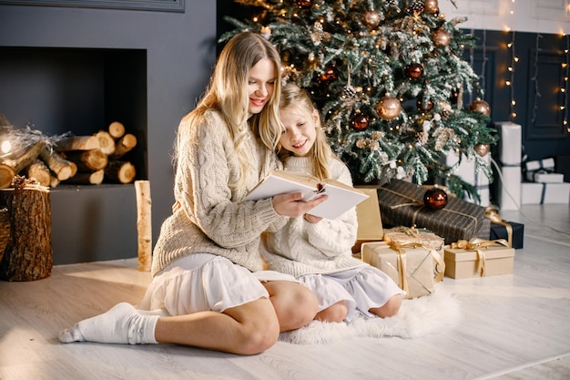 Jovem mãe e menina sentada na árvore de natal em casa e lendo um livro