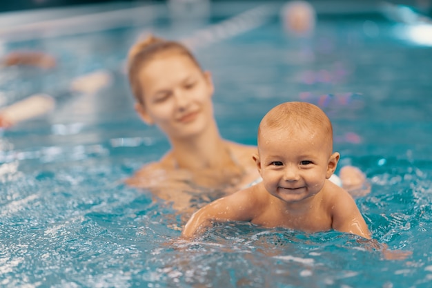 Jovem mãe e seu bebê, desfrutando de uma aula de natação do bebê na piscina. Criança se divertindo na água com a mãe