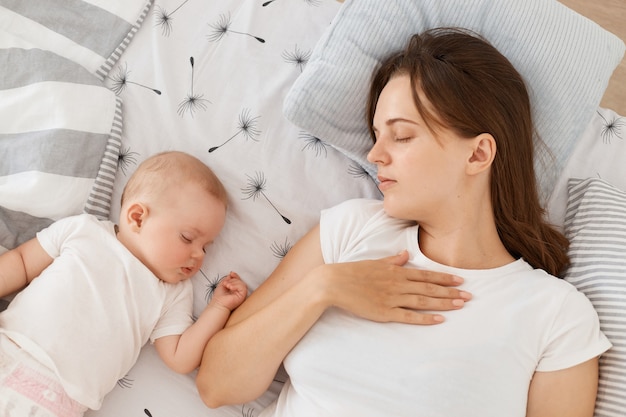 Jovem mãe e seu bebê dormindo juntos na cama, mantém os olhos fechados, vestindo camiseta branca estilo casual, deitado no travesseiro, mãe cansada descansando enquanto sua filha fofa dorme.