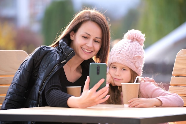 Jovem mãe e sua filha tirando foto com câmera selfie de telefone sentado no café de rua com bebidas quentes no dia ensolarado de outono Presença de mídia social no conceito de vida cotidiana