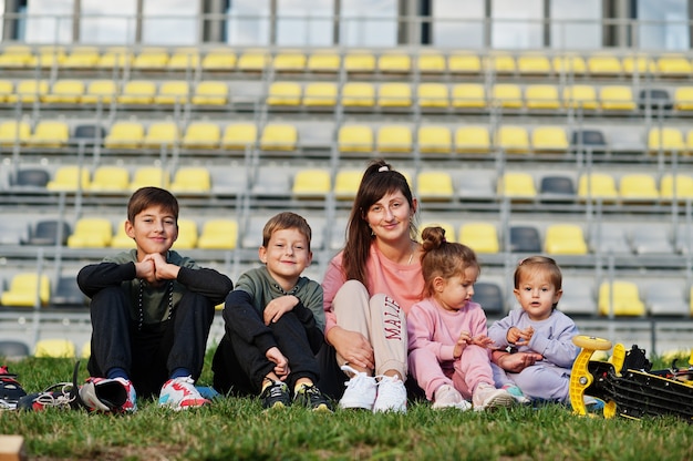 Jovem mãe elegante com quatro filhos sentados na grama contra o estádio. A família de esportes passa o tempo livre ao ar livre com patinetes e patins.