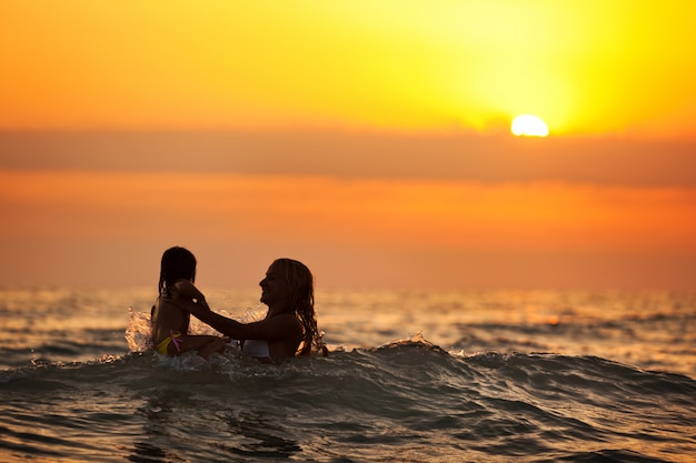 Jovem mãe em pé na água e brincando com a filha ao pôr do sol