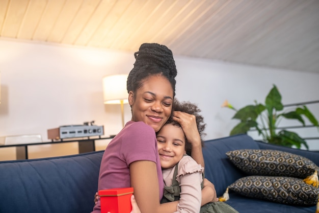 jovem mãe feliz, de pele escura, com uma filhinha fofa e um presente abraçando com os olhos fechados