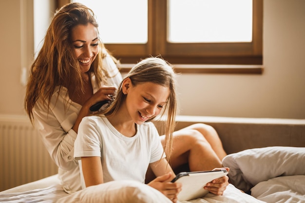 Jovem mãe feliz sorridente está penteando o cabelo da filha na cama enquanto a garota está olhando para o tablet digital.