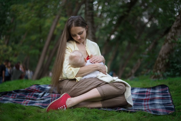 Jovem mãe linda abraçando recém-nascido e amamenta. Piquenique com bebê no parque